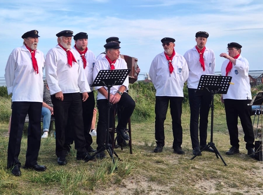 Das Foto zeigt 8 Mitglieder des Juister Shanty-Chors in weißen Hemden, roten Halstüchern, dunklen Kappen und Hosen auf der Düne am Wasserturm (Der Akkordeonspieler ist verdeckt)